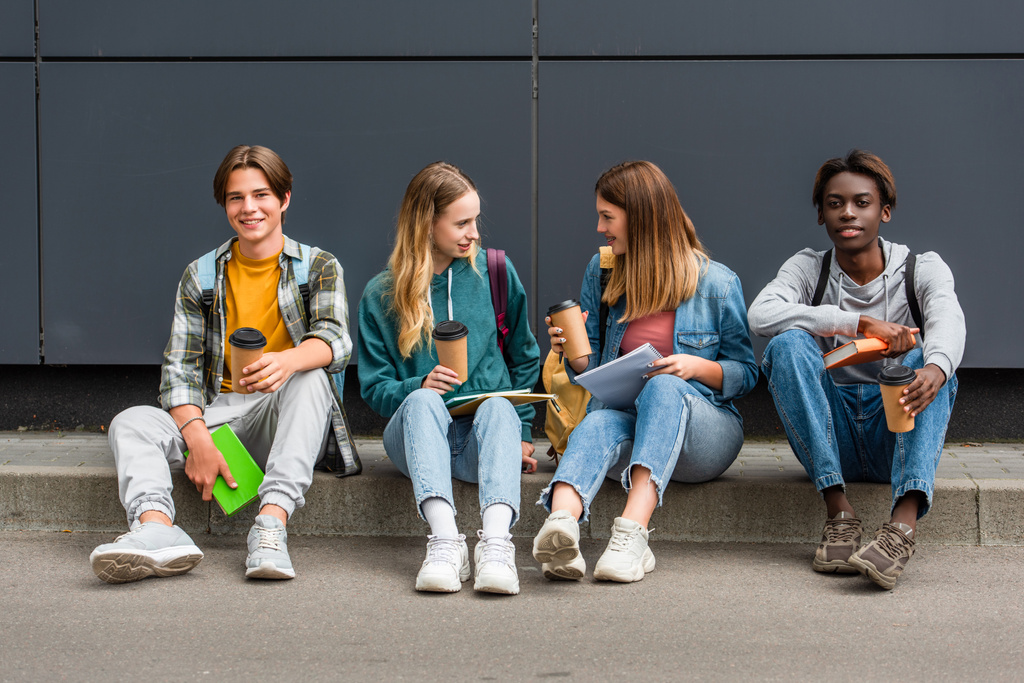 Улыбающиеся мультиэтнические подростки с кофе, чтобы пойти и рюкзаки говорить рядом со зданием  - Фото, изображение