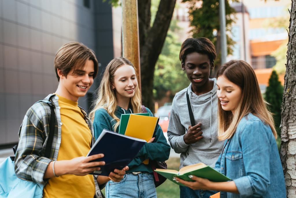 Foco seletivo de adolescentes multiétnicos positivos olhando para livro perto de árvore ao ar livre  - Foto, Imagem