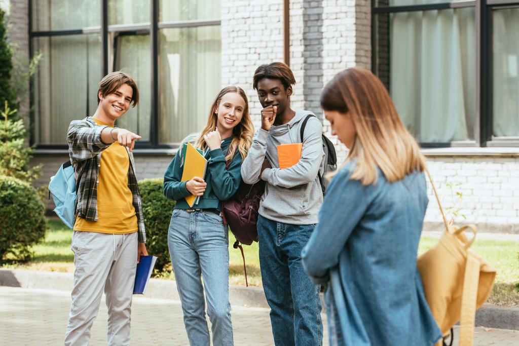 Enfoque selectivo de adolescentes multiculturales sonrientes con mochilas apuntando a una chica triste al aire libre - Foto, imagen