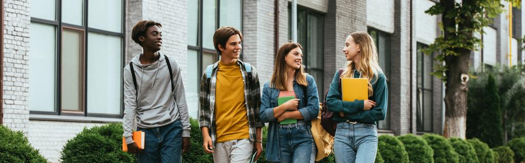 Panoráma felvétel mosolygós multikulturális tinédzser barátok jegyzetfüzetekkel séta a városi utcán  - Fotó, kép