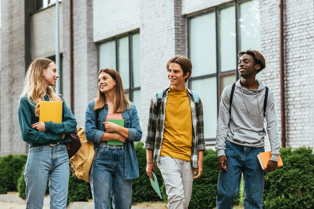 Adolescentes multiétnicos olhando para amigo com notebook enquanto caminham na rua urbana  - Foto, Imagem
