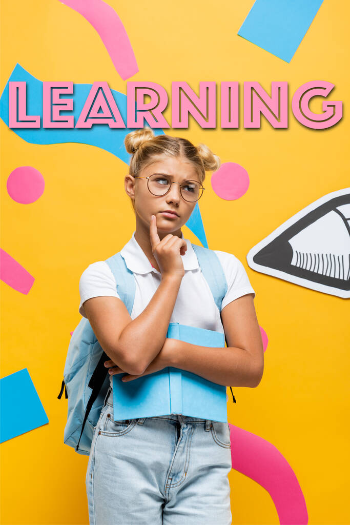 вдумчивая школьница в очках смотрит в сторону, держа книгу возле бумажного карандаша, красочных элементов и обучения буквы на желтом - Фото, изображение