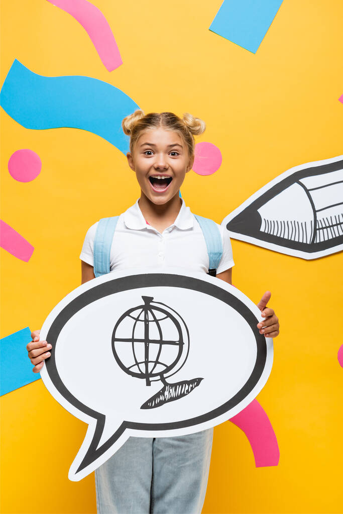 aufgeregtes Schulkind hält Sprechblase mit Globus-Abbildung in der Nähe von Papierelementen und Bleistift auf Gelb  - Foto, Bild