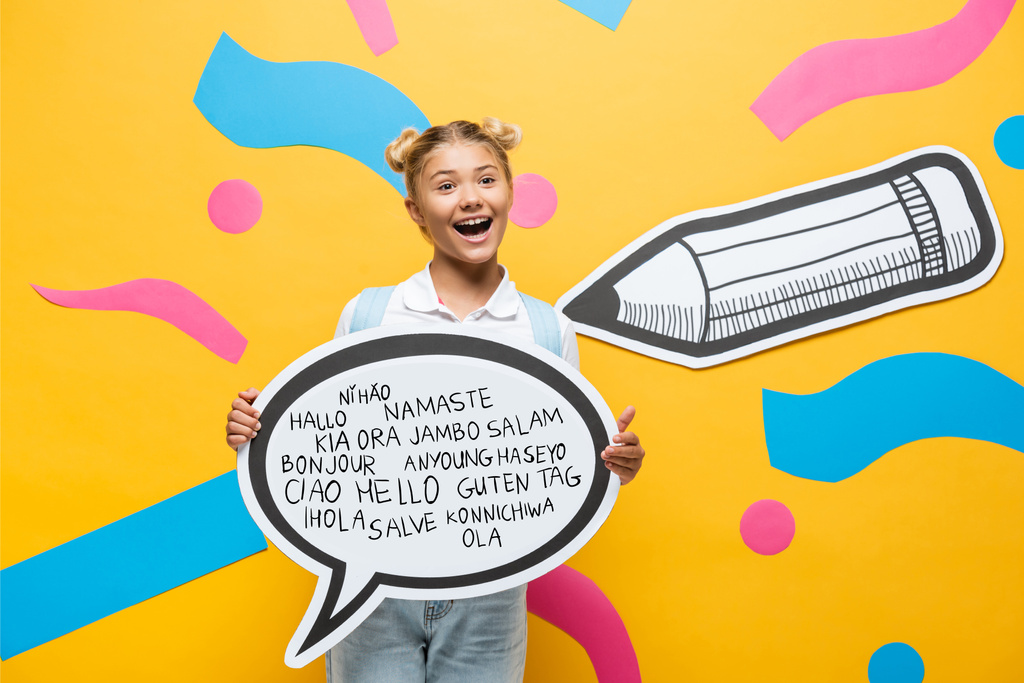 excited schoolchild holding speech bubble with greeting lettering near multicolored paper elements and pencil on yellow  - Photo, Image