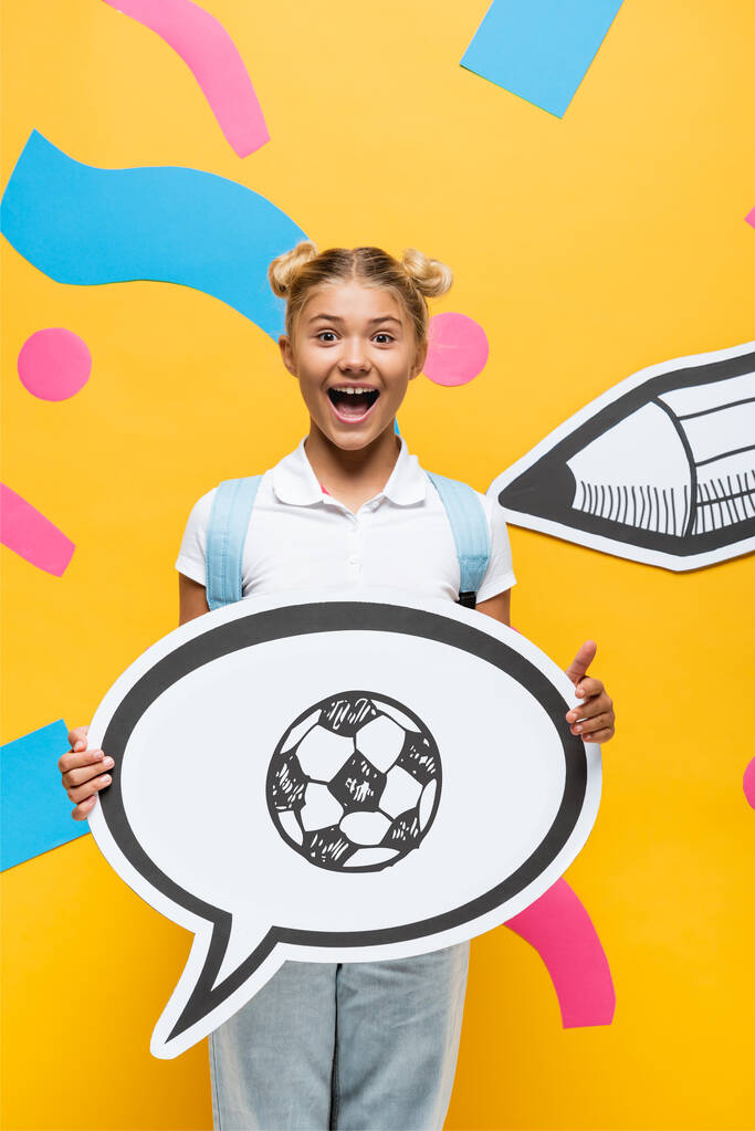 aufgeregtes Schulkind hält Sprechblase mit Fußball-Illustration in der Nähe von Papierelementen und Bleistift auf Gelb  - Foto, Bild