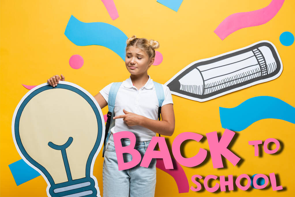 Zmatená školačka ukazující prstem na papírovou žárovku v blízkosti dekorativních prvků a zpět do školy písmo na žluté  - Fotografie, Obrázek