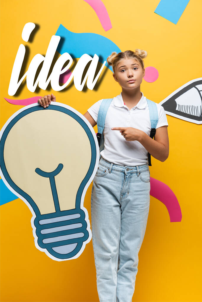 Schoolgirl with backpack pointing with finger at decorative light bulb near paper art and idea lettering on yellow background - Photo, Image