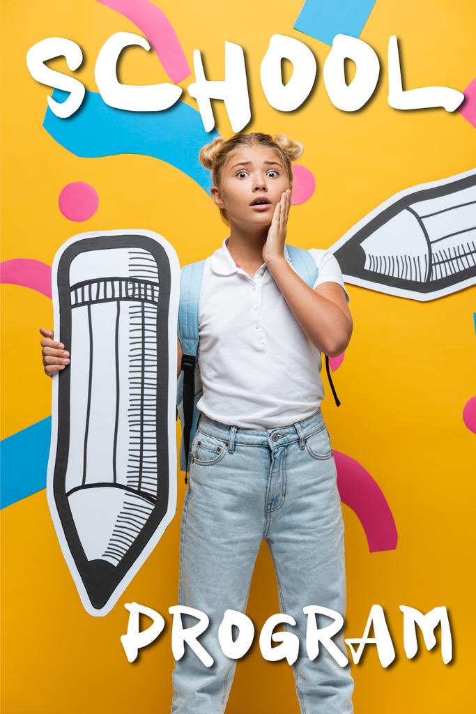 Bezorgd schoolkind met rugzak en papier potlood op zoek naar camera in de buurt van schoolprogramma belettering op geel  - Foto, afbeelding