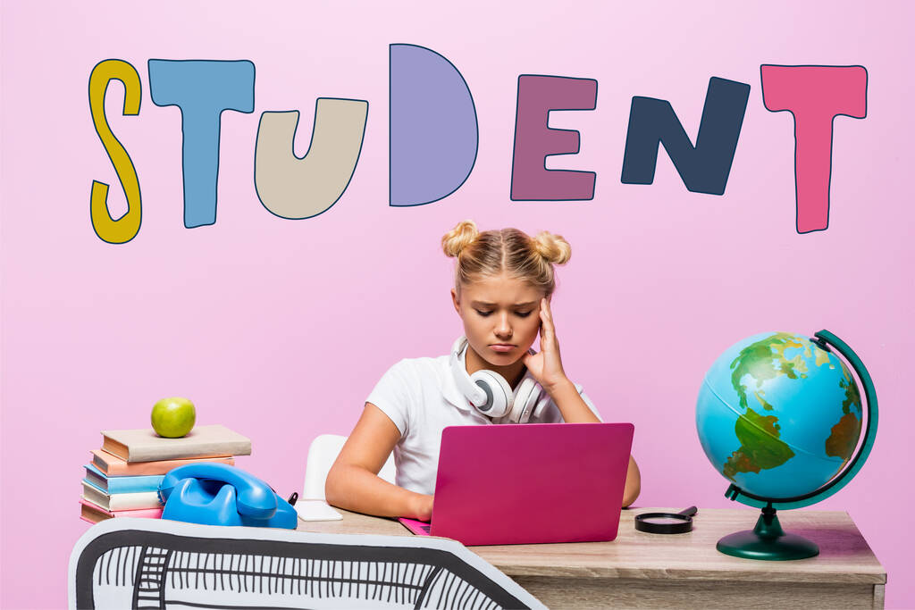 Sad kid in headphones using laptop near globe, books, retro telephone, student lettering and paper art on pink  - Photo, Image