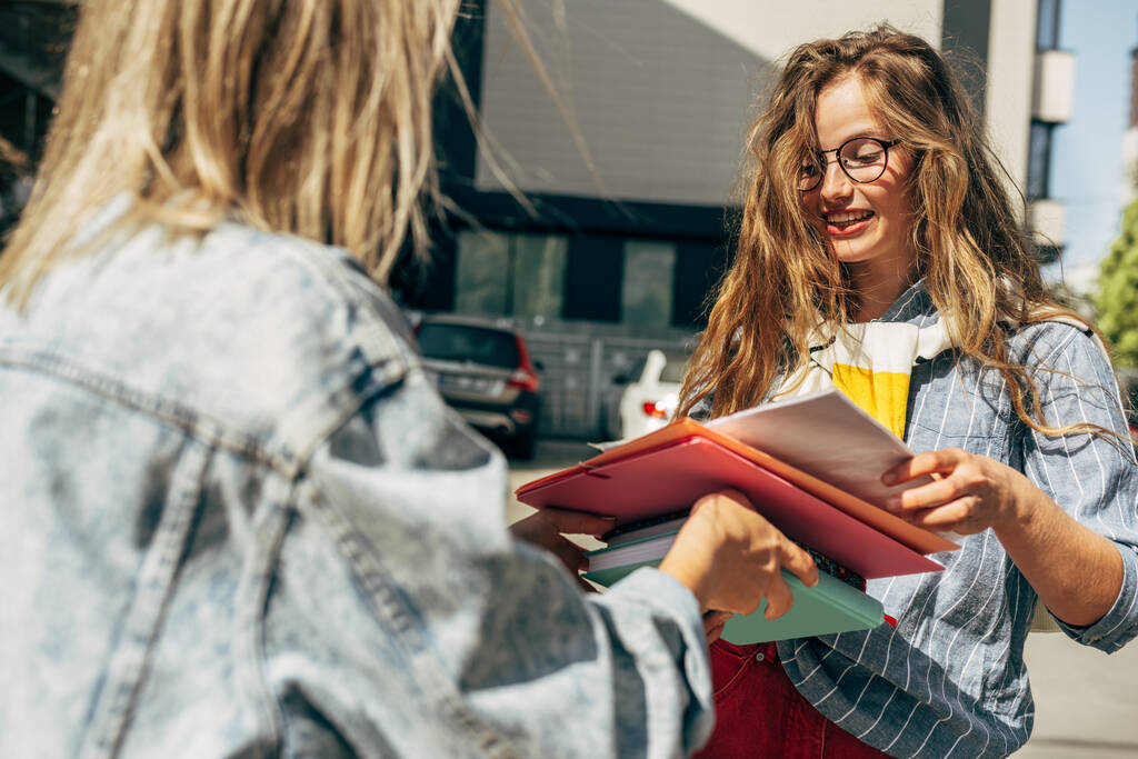 Kültéri kép két diákról, akik osztoznak a könyveken, hogy másokat tanítsanak az egyetemen. Egyetem fiatal nők főiskolai hallgatók ad a mappák és könyvek a város utcáján. - Fotó, kép