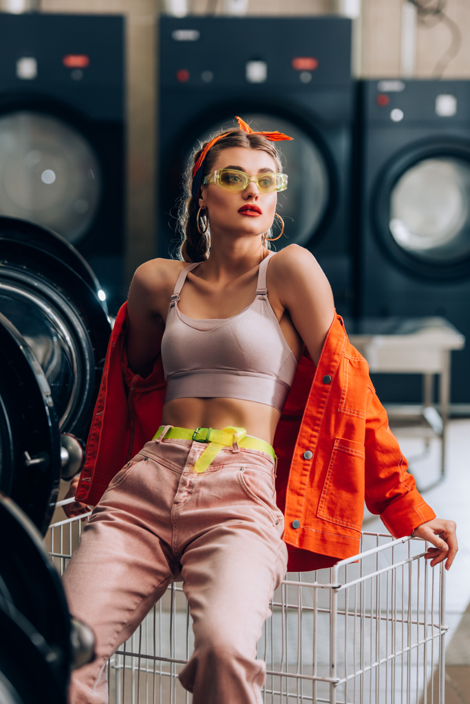 stylish woman in sunglasses looking away near basket and washing machines in laundromat - Photo, Image