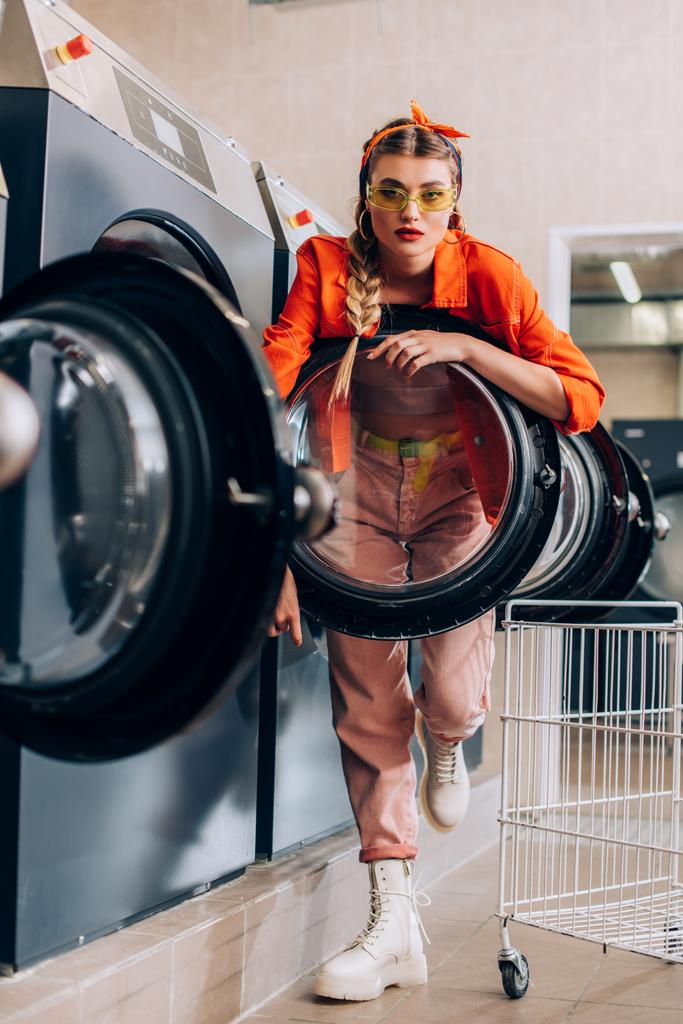 selectieve focus van stijlvolle vrouw in zonnebril kijken naar camera in de buurt van wasmachines in wasserette - Foto, afbeelding
