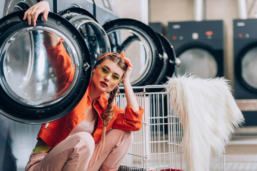mujer de moda en gafas de sol sentada cerca de carro metálico y lavadoras en lavandería - Foto, imagen
