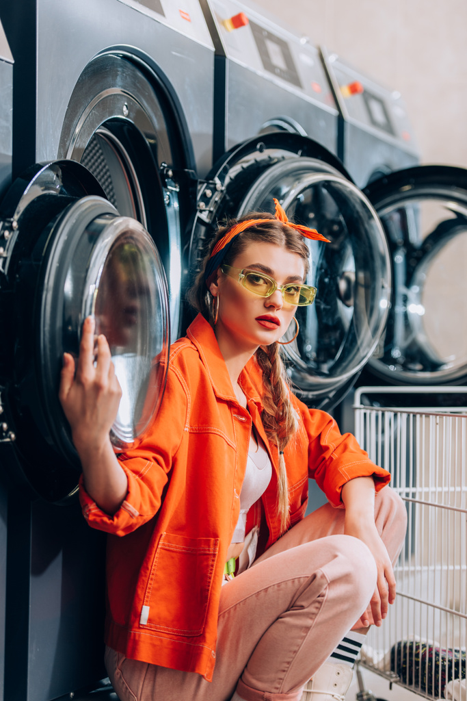 donna alla moda in occhiali da sole seduto vicino al carrello metallico e lavatrici in lavanderia - Foto, immagini