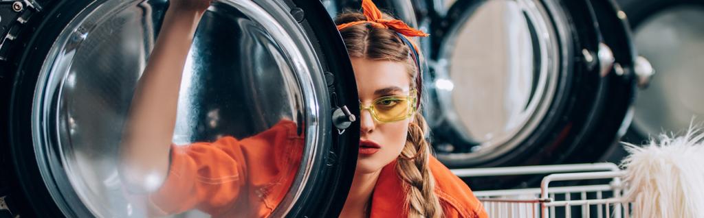 młoda kobieta w okularach przeciwsłonecznych patrząc na kamerę przez drzwi pralki, baner - Zdjęcie, obraz