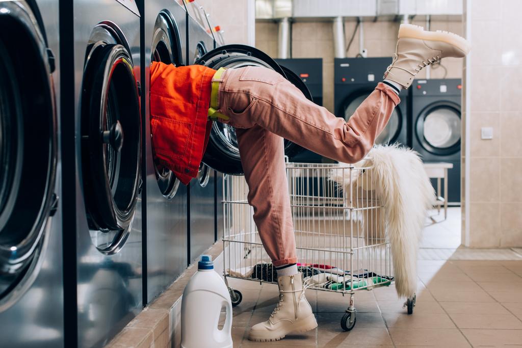 młoda kobieta sprawdzanie wewnątrz pralki w pobliżu butelki detergentu i wózka z brudnej odzieży  - Zdjęcie, obraz