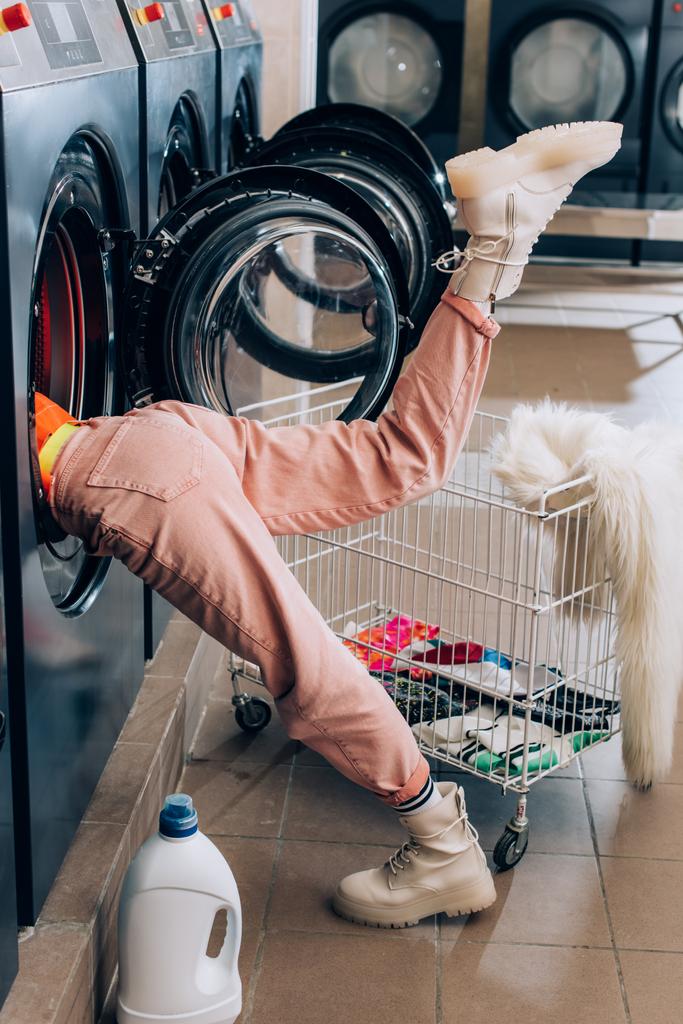 Neugierige junge Frau checkt Waschmaschine in der Nähe von Waschmittelflasche und Einkaufswagen mit schmutziger Kleidung  - Foto, Bild