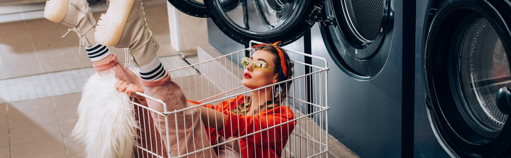 стильная женщина сидит в тележке возле стиральных машин в прачечной, баннер - Фото, изображение