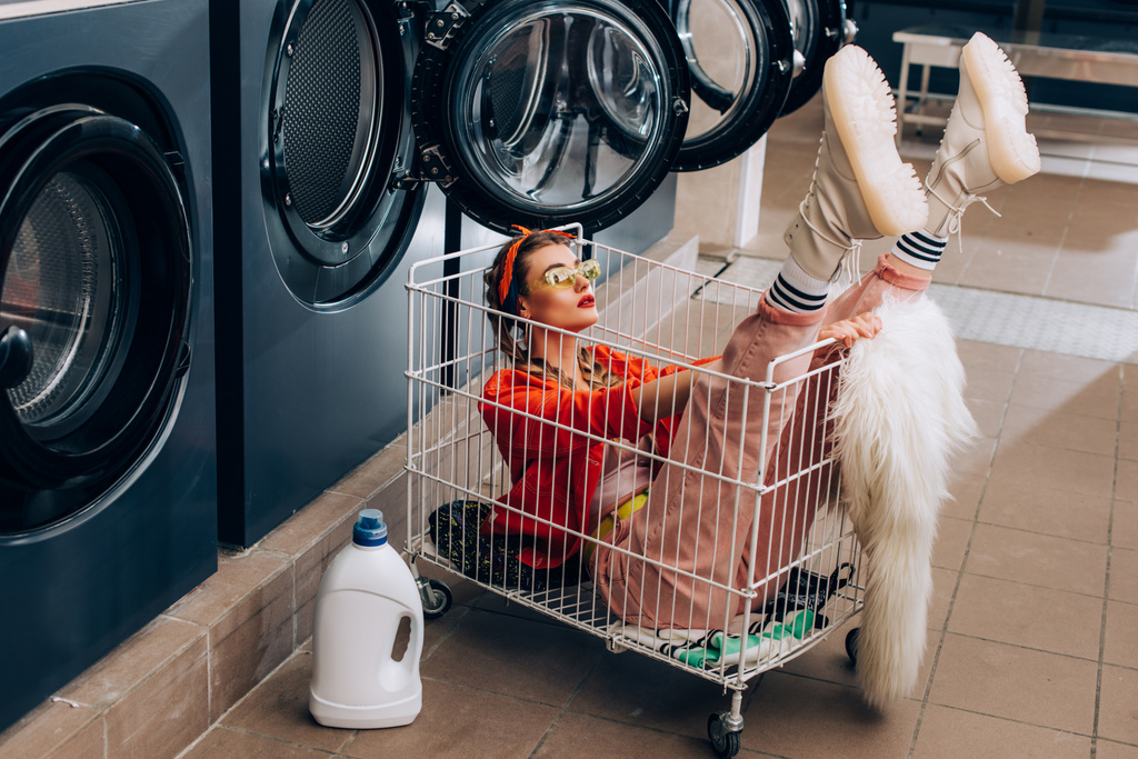 stylowa kobieta siedzi w koszyku obok pralek i butelki z detergentem w pralni - Zdjęcie, obraz
