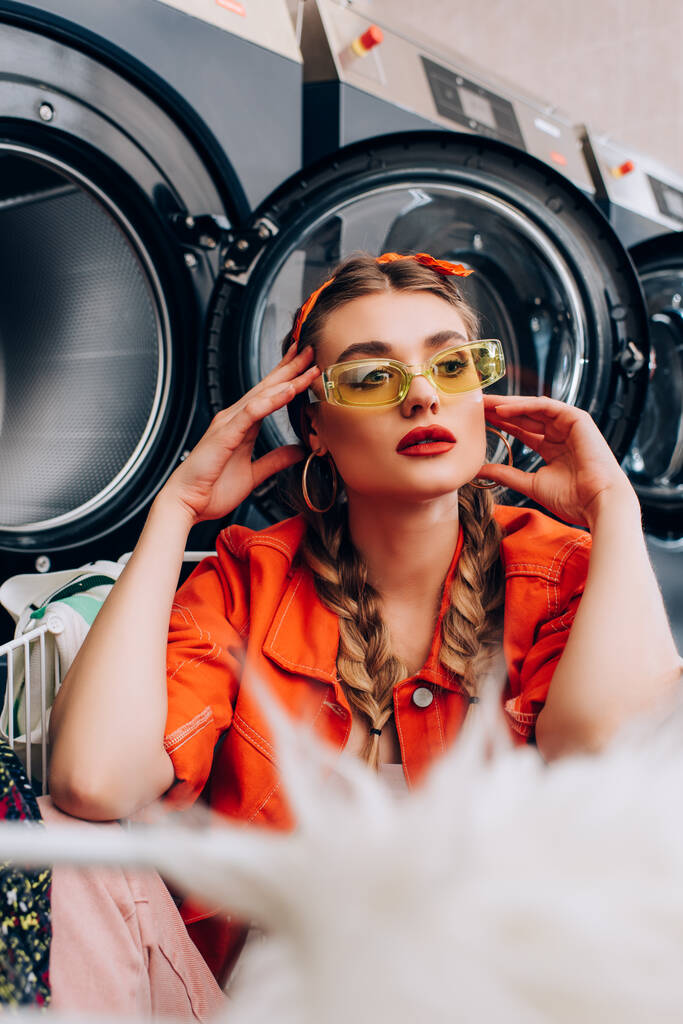 stylish woman touching face and sitting in cart near washing machines in laundromat with blurred foreground - Photo, Image