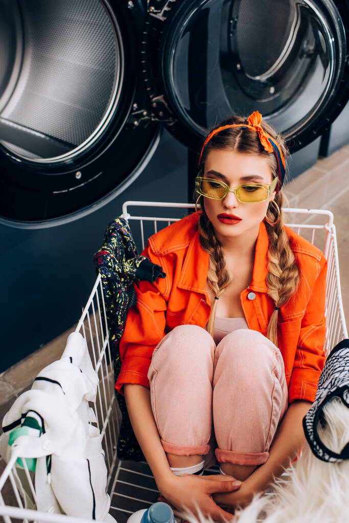 Stilvolle Frau sitzt mit Kleidung in Wäschewagen neben Waschmaschinen im Waschsalon - Foto, Bild