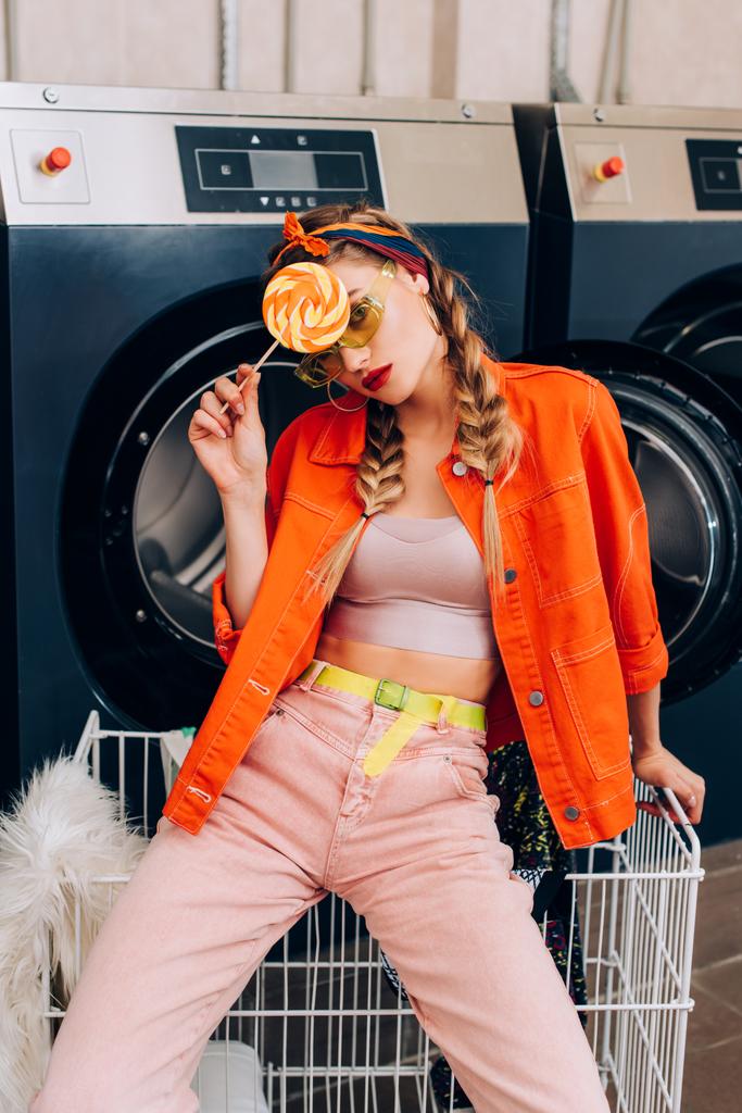 Trendfrau mit Sonnenbrille hält Lutscher neben Einkaufswagen und Waschmaschinen im Waschsalon - Foto, Bild