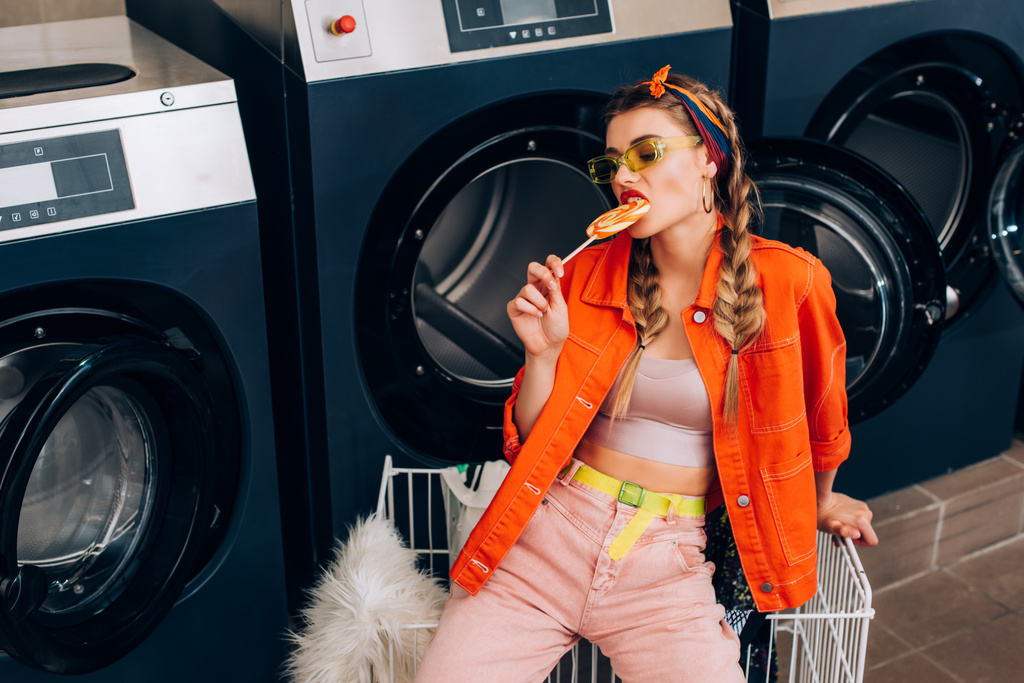 mujer de moda en gafas de sol mordiendo piruleta cerca del carro y lavadoras en lavandería - Foto, imagen