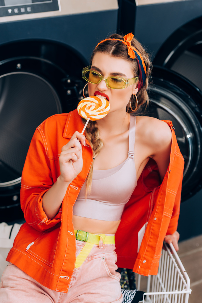 trendige junge Frau mit Sonnenbrille beißt Lutscher neben Einkaufswagen und Waschmaschinen im Waschsalon - Foto, Bild