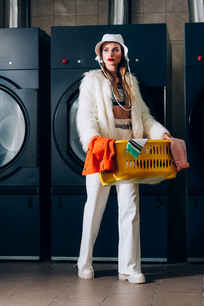 стильна молода жінка в штучній шубці і капелюсі тримає кошик з білизною біля пральних машин в пральні
 - Фото, зображення