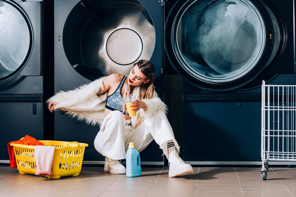 junge Frau in Kunstpelzjacke hält Plastikbecher mit Orangensaft neben Korb mit Wäsche, Waschmittelflasche und Waschmaschinen im Waschsalon - Foto, Bild