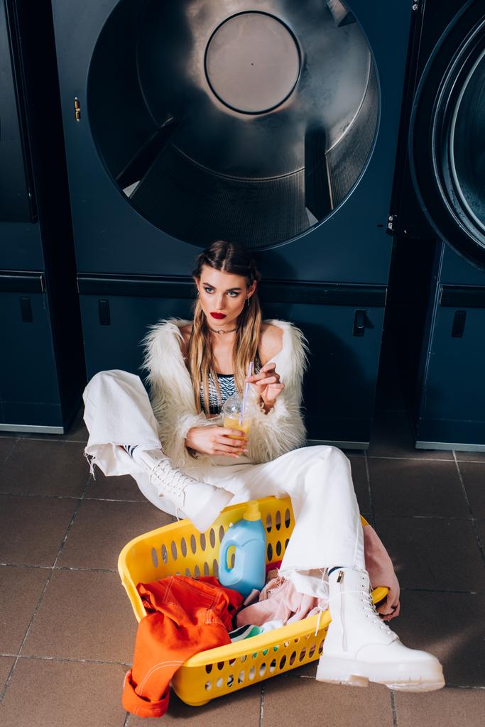 young woman in faux fur jacket sitting with orange juice near basket with clothing in laundromat - Photo, Image