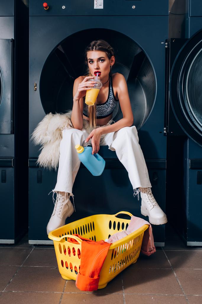 stylische Frau sitzt in Waschmaschine neben Kunstpelzjacke und trinkt Orangensaft neben Korb mit schmutziger Kleidung im Waschsalon - Foto, Bild