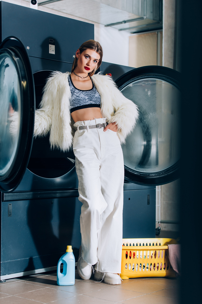 mujer joven y elegante en chaqueta de piel sintética de pie con la mano en el bolsillo en la lavandería pública - Foto, Imagen