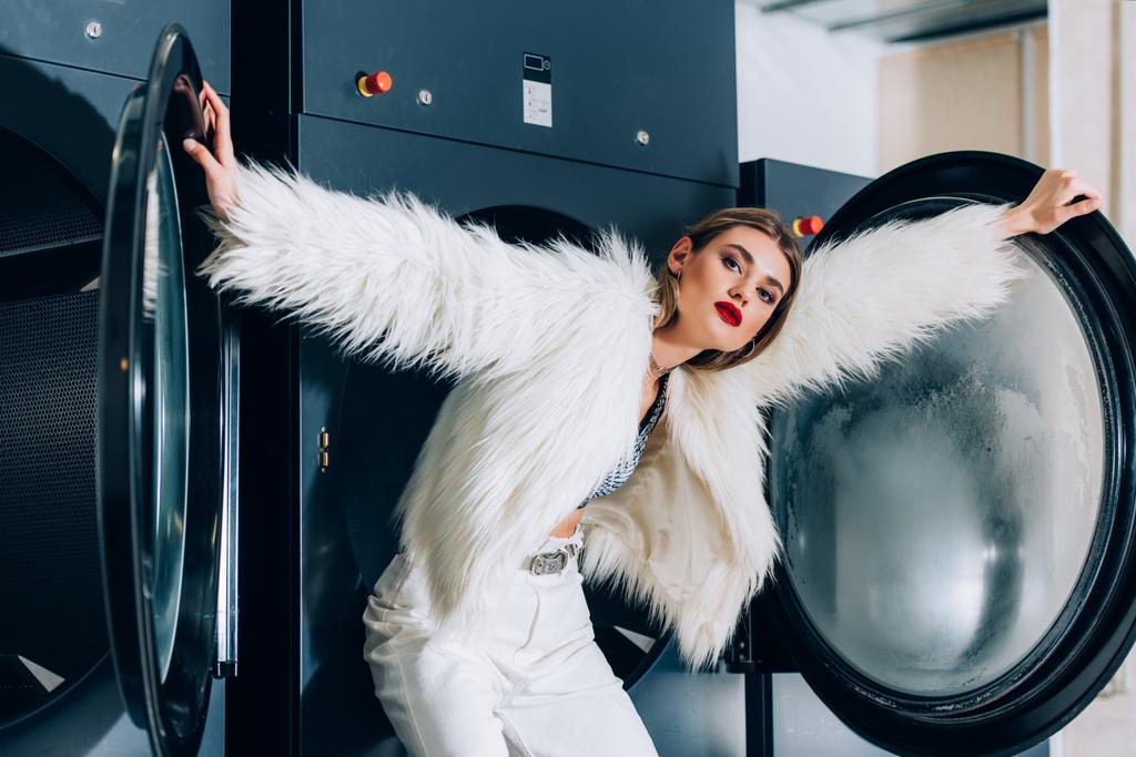 Çamaşırhanedeki çamaşır makinelerinin yanında duran, sahte kürk ceketli, modaya uygun genç bir kadın. - Fotoğraf, Görsel