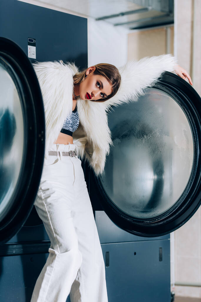 giovane donna elegante in pelliccia sintetica giacca in piedi vicino lavatrici in lavanderia pubblica - Foto, immagini