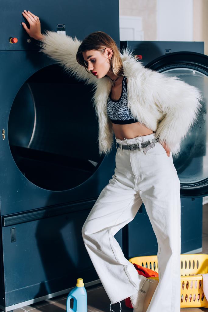jonge stijlvolle vrouw in faux fur jas leunend op wasmachine in openbare wasserette - Foto, afbeelding