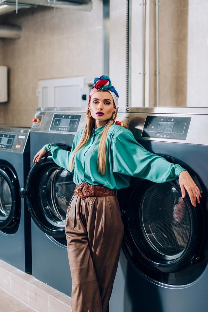 stijlvolle vrouw in tulband staan in de buurt van wasmachines in wasserette  - Foto, afbeelding