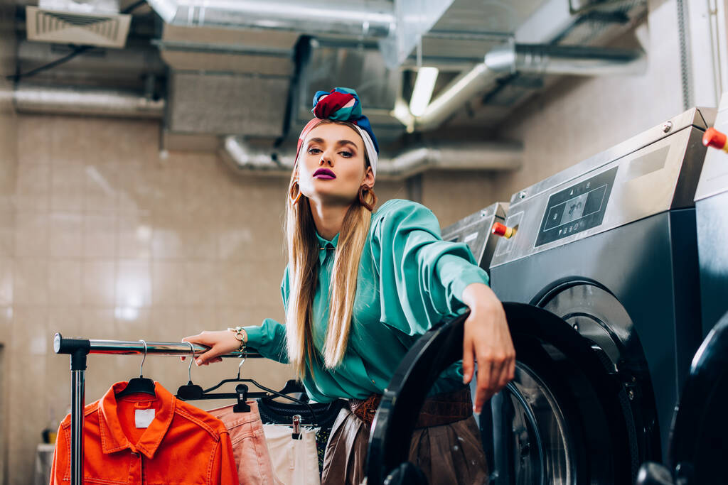 mulher na moda em turbante de pé perto de rack de roupas e máquinas de lavar roupa na lavanderia moderna  - Foto, Imagem