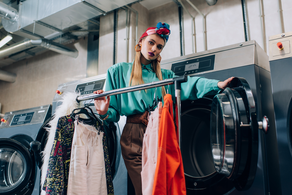 junge und stylische Frau im Turban steht neben Kleiderständern und Waschmaschinen im modernen Waschsalon  - Foto, Bild