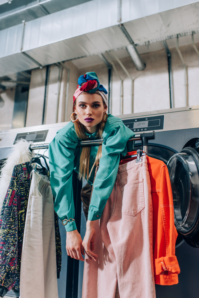 молодая женщина в тюрбане опирается на вешалку для одежды и стиральные машины в прачечной  - Фото, изображение