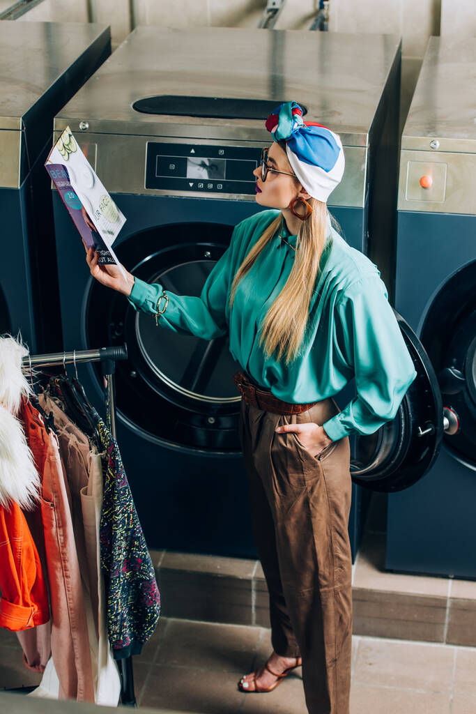 mujer con estilo en turbante y gafas de lectura revista y de pie con la mano en el bolsillo en la lavandería  - Foto, imagen