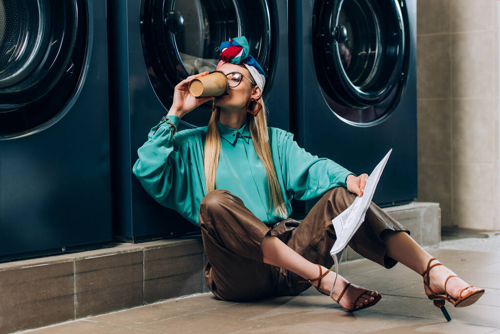 модна жінка в окулярах і турбан тримає газету і п'є каву в пральні
  - Фото, зображення