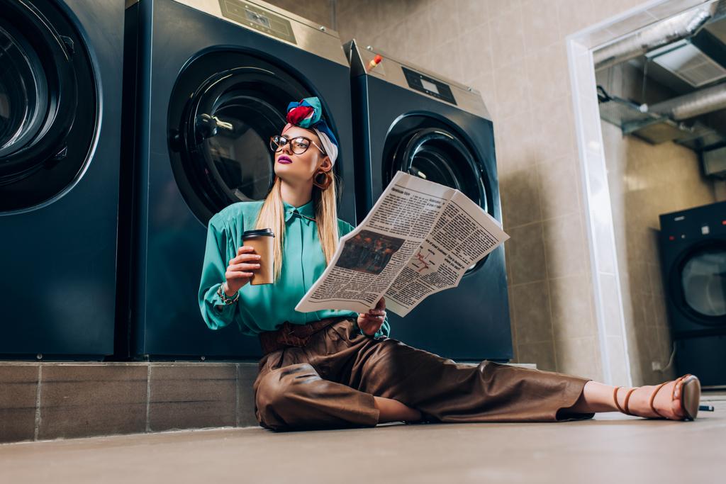 modna kobieta w okularach i turban trzymając papierową filiżankę i gazetę podczas siedzenia na podłodze w pralni  - Zdjęcie, obraz