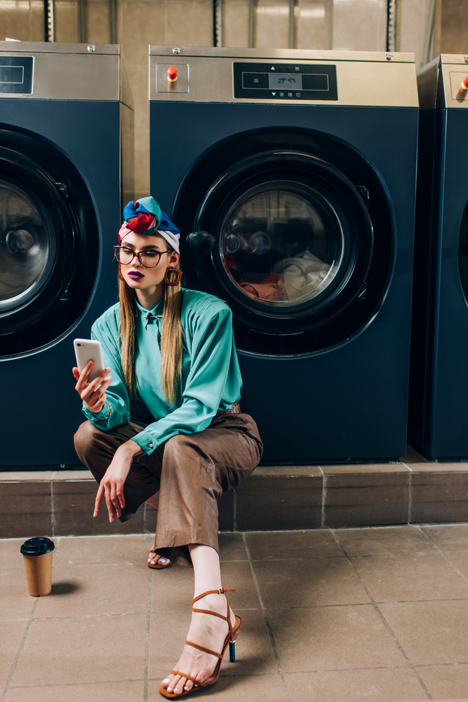 mujer joven en gafas y turbante sosteniendo teléfono inteligente cerca de taza de papel en lavandería  - Foto, imagen