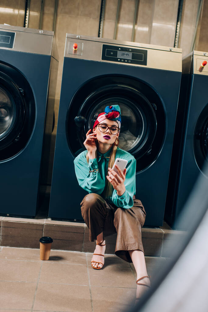 mujer joven en gafas y turbante mirando teléfono inteligente cerca de taza de papel en lavandería  - Foto, Imagen