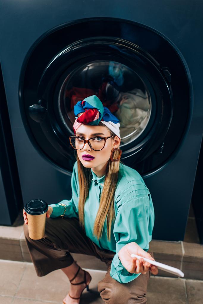Tiefansicht einer jungen Frau mit Brille und Turban, die Smartphone und Pappbecher im Waschsalon hält  - Foto, Bild