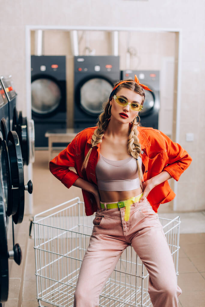 stylish woman in headband and sunglasses posing with hands on hips in modern laundromat  - Photo, Image