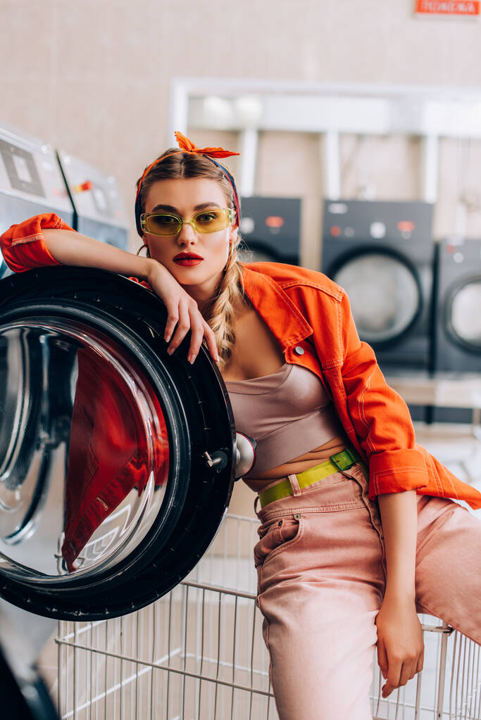 młoda i stylowa kobieta w opasce i okularach przeciwsłonecznych w nowoczesnej pralni  - Zdjęcie, obraz
