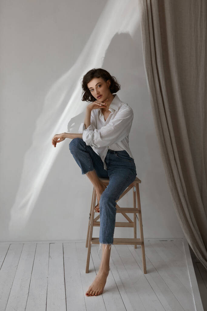 Žena středního věku s štíhlým tělem v bílé košili a modré džíny při pohledu do kamery a pózování na stolici uvnitř - Fotografie, Obrázek