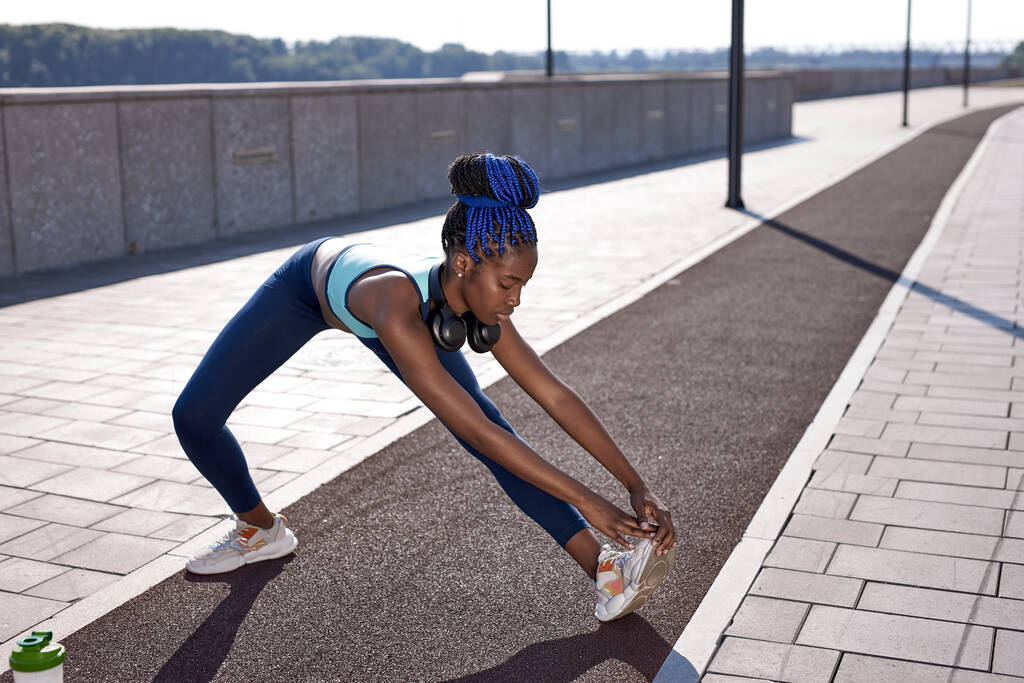 atleet afro vrouw stretching hamstring beenspieren tijdens outdoor hardlopen workout - Foto, afbeelding